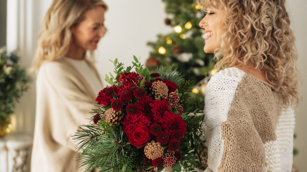 Ragazza che tiene in mano un boquuet natalizio di fiori rossi con in sfondo un albero di Natale e una donna che lo sistema.