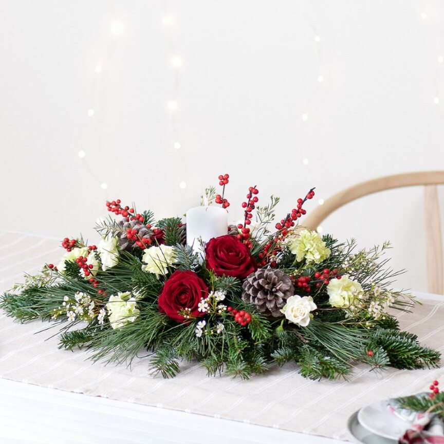 Centrotavola di Natale elegante con una candela al centro di rose rosse, fiori gialli e bianche, pigne e del verde.